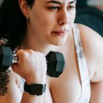 Strength Equipment - Concentrated woman in sportswear sitting on bench and lifting dumbbell during weight loss workout in gym with fitness equipment