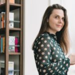 Online Bookstores - Side view of positive young female student with long dark hair in elegant dress using smartphone and smiling while standing near window in light library