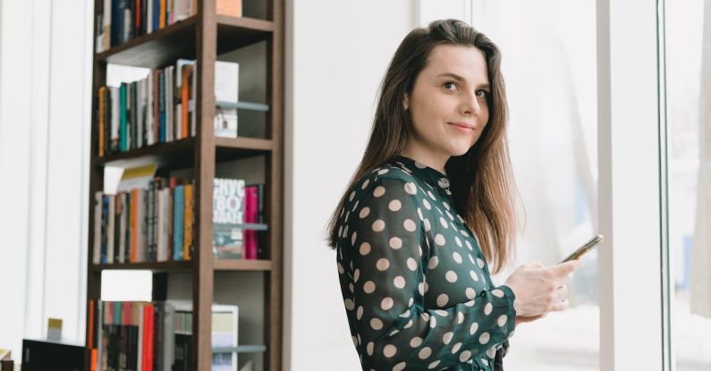 Online Bookstores - Side view of positive young female student with long dark hair in elegant dress using smartphone and smiling while standing near window in light library