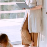 Online Toy Stores - Adult female wearing casual clothes standing in corner of room and using laptop while spending time with daughter in daytime