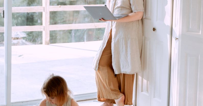 Online Toy Stores - Adult female wearing casual clothes standing in corner of room and using laptop while spending time with daughter in daytime
