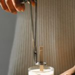 DIY Workspace - Unrecognizable African American female standing at wooden table with candle and steel wick dipper in light room with sunlight