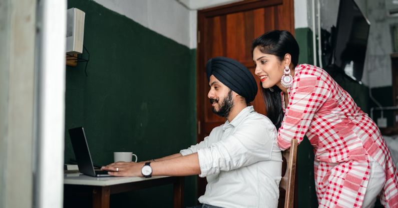 Home Office - Side view of positive young Sikh man in shirt and turban working on laptop at home while wife leaning on chair behind