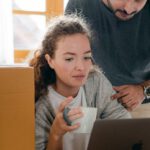 Black Friday - Thoughtful woman surfing internet on netbook while sitting with cup of hot drink near ethnic boyfriend in watch and casual wear standing near cardboard boxes after moving into new flat