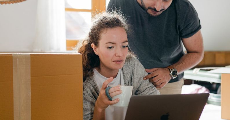 Black Friday - Thoughtful woman surfing internet on netbook while sitting with cup of hot drink near ethnic boyfriend in watch and casual wear standing near cardboard boxes after moving into new flat